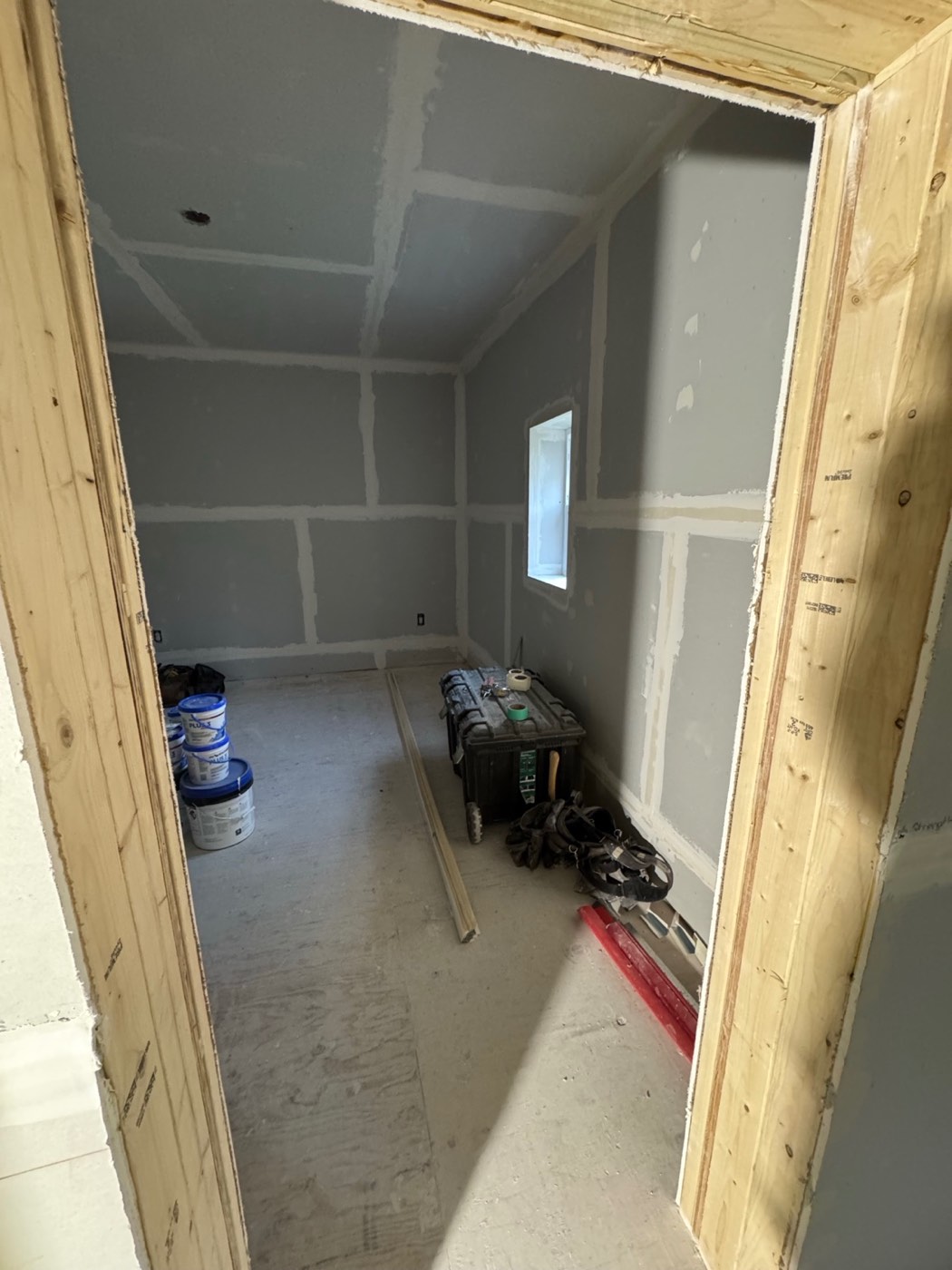 The same bedroom seen through the framed doorway, with taped drywall under the sloped ceiling and the window on the right