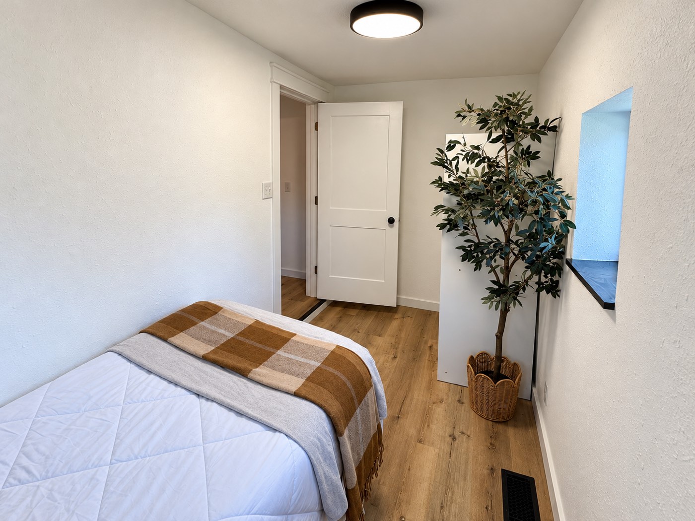 Finished narrow bedroom with a white panel door, plank flooring, white trim and a recessed window with a black sill