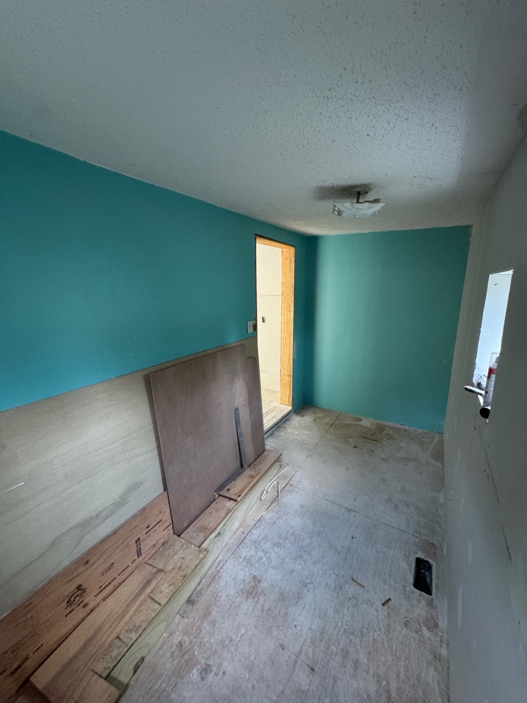 Narrow bedroom during tear-out, with teal walls, plywood leaning on the wall and the subfloor exposed