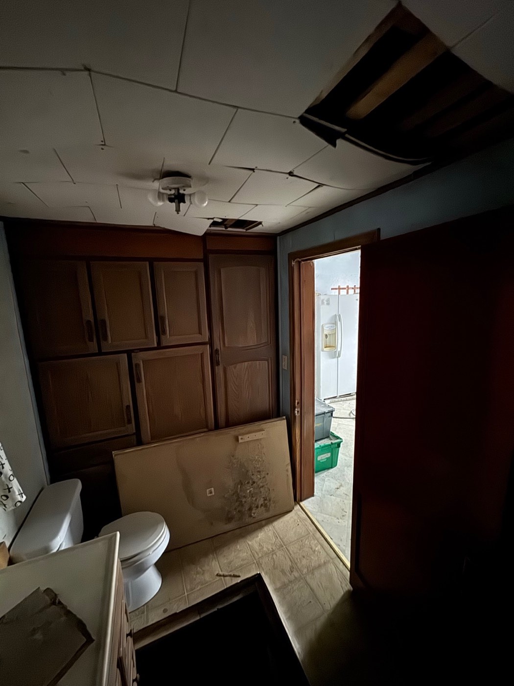 Original bathroom looking toward the door, with built-in wood cabinets, a toilet and a damaged ceiling