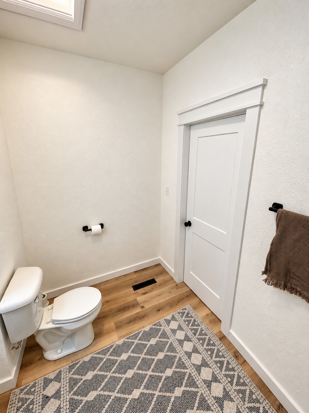 The finished bathroom looking toward the door, with white walls, a white shaker door, matte black hardware and wood-look flooring