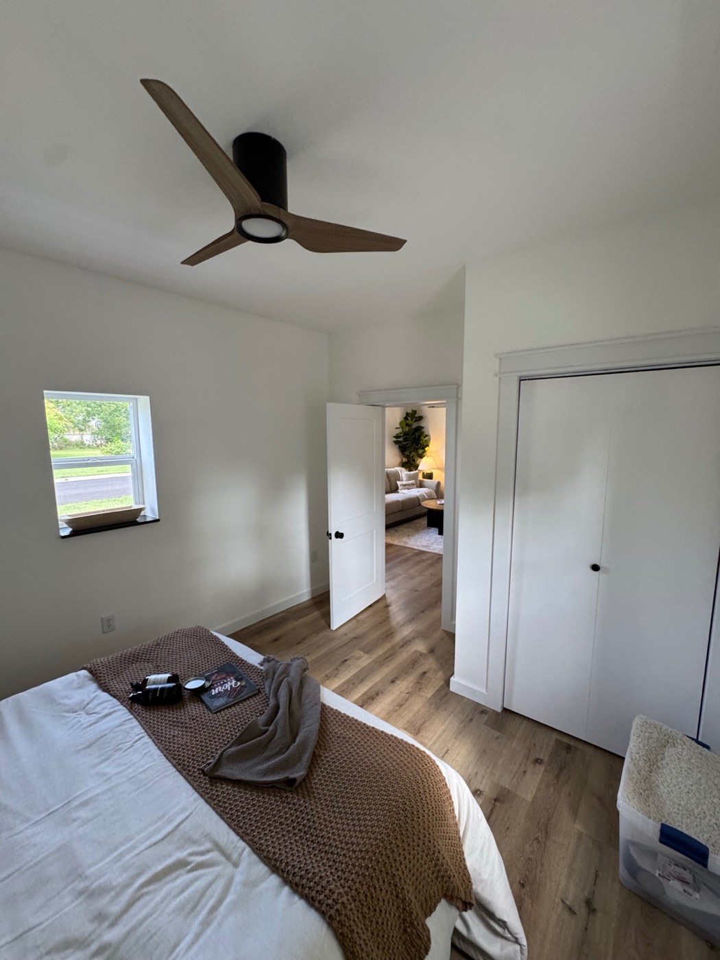 The finished bedroom from the same corner: white walls, plank flooring, a white panel door, bifold closet doors and a ceiling fan
