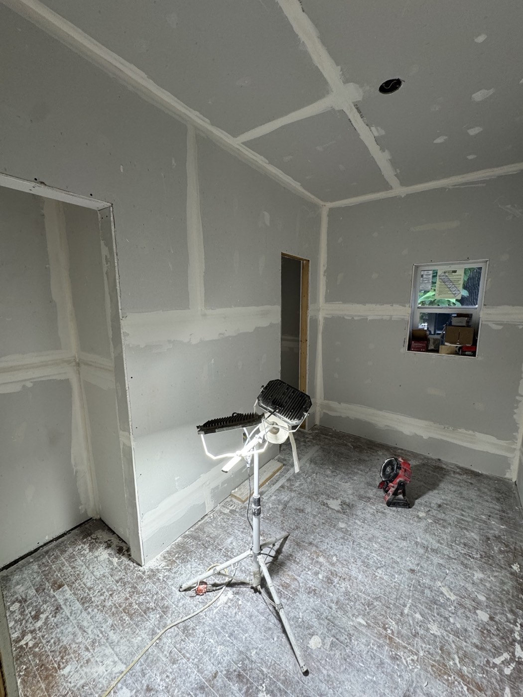Small bedroom during renovation with taped and mudded drywall, a work light, the window on the back wall and the closet opening on the left